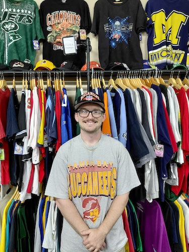 A man wearing a gray Tampa Bay Buccaneers t-shirt stands in front of a clothing rack filled with various sports jerseys and t-shirts. He is wearing a black cap and glasses.