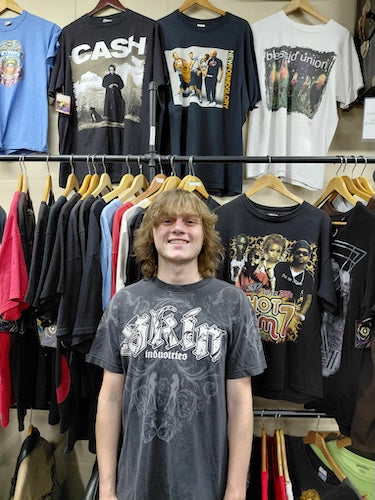 A smiling young man with long blonde hair stands in front of a clothing rack wearing a faded dark gray graphic t-shirt with 'SIREN' and 'Inanities' printed on the front. Behind him, other band t-shirts are hanging on wooden hangers on a store display.