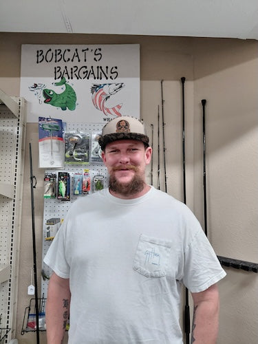 A man with a beard and a camouflage cap stands in a retail store wearing a white t-shirt with a chest pocket. Behind him is a pegboard with fishing lures, a sign reading 'BOBCAT'S BARGAINS', and fishing rods hanging on the wall.