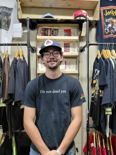 A man wearing a dark gray graphic t-shirt with the text 'I'm not dead yet' and a blue cap stands in front of a clothing rack in a retail store.